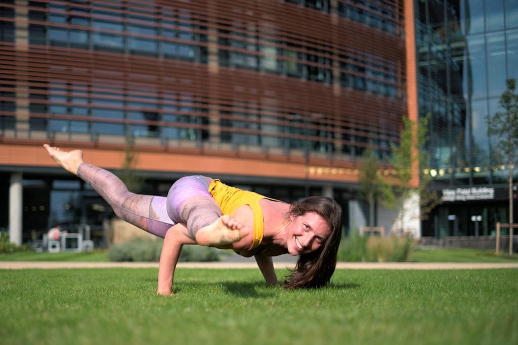 Christina Weis in a one-arm side-crow arm balance on the grass outside the De Montfort University campus